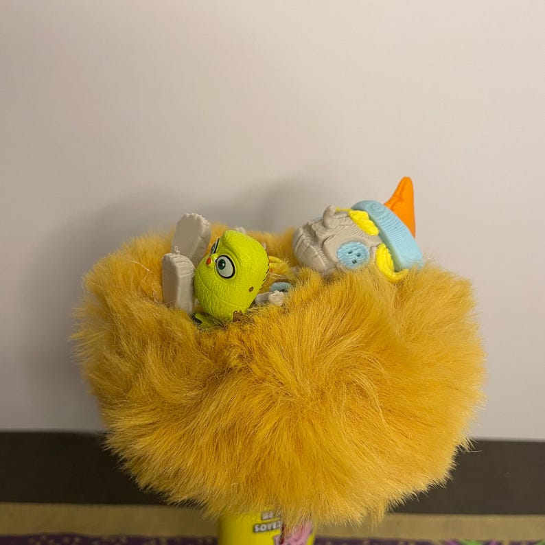 May include: A plush, yellow toy with a fuzzy texture, featuring a small, green creature with a large eye and a gray and blue toy on top. The toy has a cylindrical base with the word "SOF" visible.