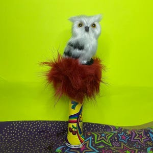 May include: A decorative owl figurine with white and black feathers and yellow eyes sits atop a red, fluffy base. The base is attached to a yellow cylinder with a rainbow design and the number 7.