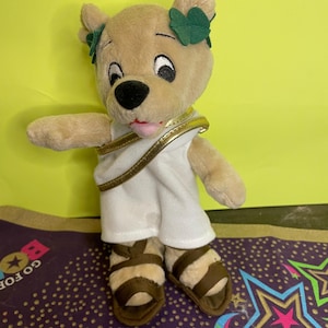 Puede incluir: Un oso de peluche marrón que lleva una túnica blanca con ribete dorado y una corona de laurel verde. El oso lleva sandalias marrones.