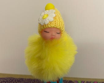 May include: A yellow pen topper shaped like a baby doll. The doll has a yellow knitted hat with a white daisy and a yellow pom-pom. The doll is wrapped in a fluffy yellow material.
