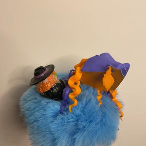 Puede incluir: Una decoraci&oacute;n de Halloween caprichosa con una figura de bruja con cabello naranja y un sombrero negro, descansando sobre una base azul esponjosa. La bruja est&aacute; adornada con un sombrero morado y rizos naranjas, creando una est&eacute;tica l&uacute;dica y festiva.