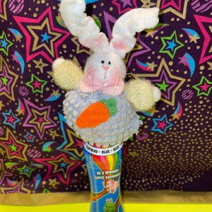 Può includere: Un giocattolo di Pasqua di peluche con un coniglietto di peluche con pelliccia bianca, dettagli rosa e una carota. Il coniglietto è appollaiato su una bottiglia blu con il testo "BE A WINNER! SOYEZ GAGNANT!" e illustrazioni di un elefante e una rana.