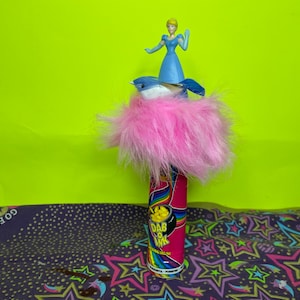May include: A pink and rainbow-colored Dab O' Ink marker is topped with a blue bird, a pink feather boa, and a Cinderella figurine. The marker has a gold pot of gold graphic and the text "Dab O' Ink".