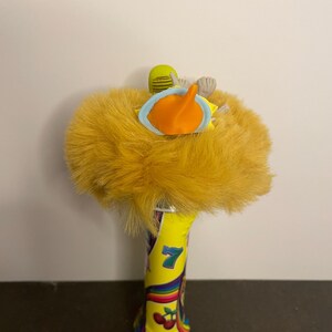 May include: A whimsical, yellow plush toy with a duck-like design. The toy features a fluffy, textured body, an orange beak, and a light blue ring. The base is a yellow cylinder with a rainbow design and the number 7.
