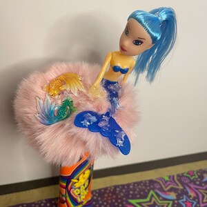 May include: A whimsical pencil topper featuring a mermaid doll with blue hair and a blue tail. The doll sits atop a fluffy pink base adorned with plastic fish. The pencil has a colorful design.