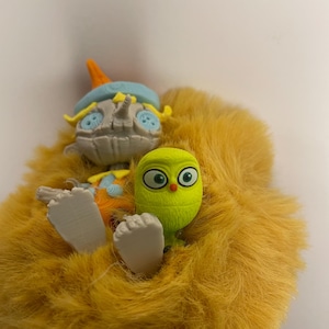 May include: A whimsical, plush toy featuring a light yellow, furry body. The toy has a gray, scarecrow-like head with a blue hat and orange accents. A small, green, owl-like figure is nestled in the fur, adding a playful touch.