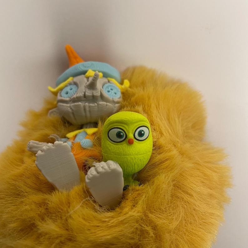 May include: A whimsical, plush toy featuring a light yellow, furry body. The toy has a gray, scarecrow-like head with a blue hat and orange accents. A small, green, owl-like figure is nestled in the fur, adding a playful touch.