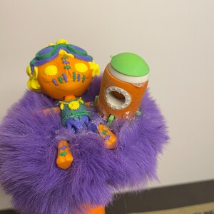 May include: A whimsical, handmade doll with an orange head, yellow hair, and green goggles. It wears a purple furry coat and holds an orange and green rocket. The doll's outfit includes a blue and green vest and orange shoes.