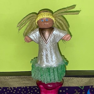 May include: A Cabbage Patch Kid doll dressed as an angel with a white dress, green glitter skirt, and yellow yarn hair. The doll has blue eyes and a brown face. The doll is wearing a gold halo and has the letters "CPK" on its chest.