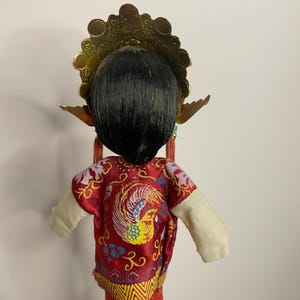 May include: A traditional Chinese doll with black hair and a gold headdress. The doll wears a red jacket with a phoenix design and white sleeves. The doll's costume is detailed with gold and yellow accents.