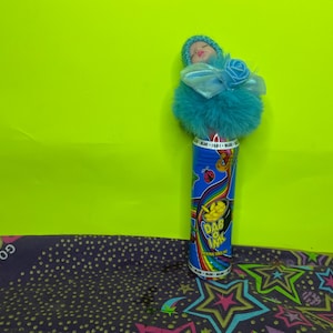 May include: A blue and red "Dab O Ink" container with a baby doll topper. The container has a rainbow design and the words "Dab O Ink" in yellow. The baby doll has a blue hat and a blue bow.