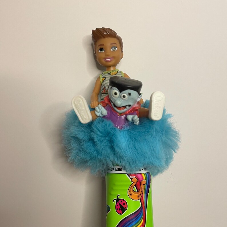 May include: A doll of a young man and a cartoon vampire are seated on a fluffy blue object, atop a green can with a rainbow design and a ladybug. The young man doll has brown hair and is wearing a yellow and white patterned shirt.