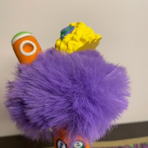 May include: A whimsical, colorful toy with a fluffy purple center. It features an orange cylindrical element with a white circle and a green stripe, and a yellow, textured top with blue and green accents.