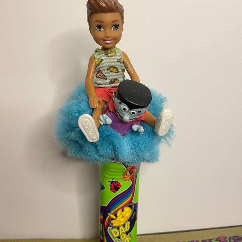 May include: A doll of a young boy wearing a taco-print tank top and red shorts sits atop a fluffy blue cushion. A small monster doll with a black hat and purple shirt is also on the cushion. The dolls are on a green and yellow container with the text "Dab Dok".