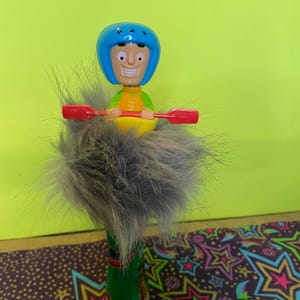 Puede incluir: Una figurita de juguete de una persona con un casco azul y un chaleco salvavidas amarillo, sosteniendo un remo rojo y amarillo, sentada en un kayak amarillo. La figurita está encima de un pompón gris y esponjoso.