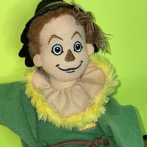 May include: A plush toy of the Scarecrow from the Wizard of Oz. The toy is wearing a green and brown outfit with a yellow straw hat and a brown belt. The Scarecrow has a friendly face with a stitched-on smile and brown eyes.