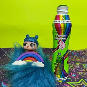 May include: A green and rainbow-colored bottle with the text "BE A WINNER! SOYEZ GAGNANT!" and a pink elephant and frog graphic. A doll with blue hair and a rainbow sits on a blue fluffy base.