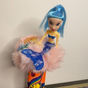 May include: A mermaid doll with blue hair and a blue tail sits atop a pink, fluffy base. The doll has a blue top and is perched on a colorful, cylindrical object with a rainbow design.