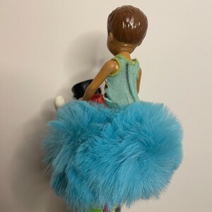 May include: A doll wearing a light blue fluffy skirt and a light green top. The doll has brown hair and is holding a black object. The doll's outfit is colorful and vibrant.
