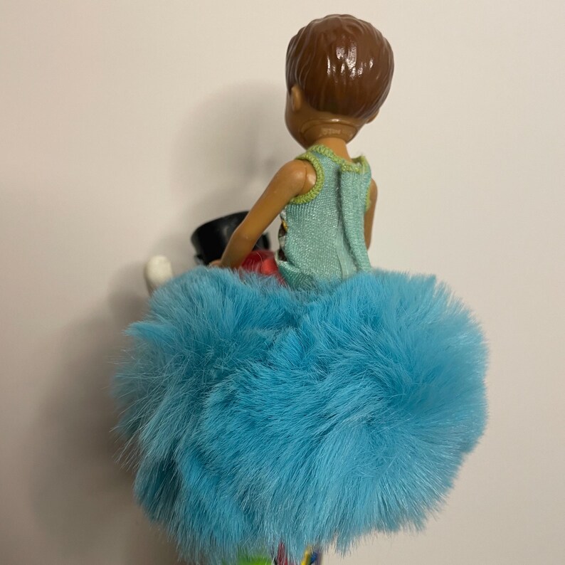May include: A doll wearing a light blue fluffy skirt and a light green top. The doll has brown hair and is holding a black object. The doll's outfit is colorful and vibrant.