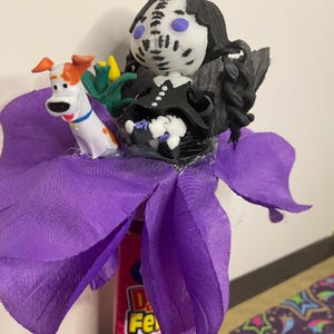 May include: A whimsical, handmade craft featuring a purple flower with a Max the Dog figurine and a doll wearing a black dress and a mask. The doll has black hair and a white face with blue eyes. The flower is made of purple fabric.