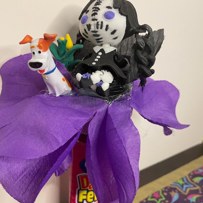 May include: A whimsical, handmade craft featuring a purple flower with a Max the Dog figurine and a doll wearing a black dress and a mask. The doll has black hair and a white face with blue eyes. The flower is made of purple fabric.