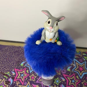 May include: A blue fuzzy decorative item topped with a cartoon rabbit figurine holding a carrot. The rabbit is gray, white, and tan. The item is likely a car accessory or a decorative piece.