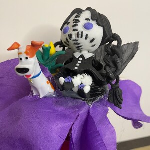 May include: A whimsical figurine featuring a doll with a white face, black hair, and a black dress, accompanied by a cartoon dog. The doll is seated on a purple fabric flower, with green and yellow accents. The dog has orange and white markings.