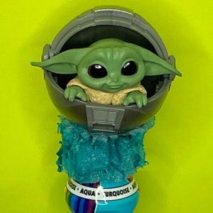 May include: A green and grey toy figure of a character from the Star Wars franchise, sitting in a grey pod. The pod is surrounded by a blue and white fluffy material. The base of the toy is a blue and white striped cylinder with the words "Aqua Turquoise" printed on it.
