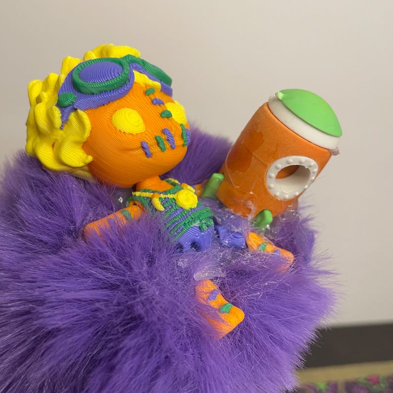 May include: A colorful 3D-printed figurine of a character with orange skin, yellow hair, and blue and green accents. The character is holding an orange and green object, all resting on a bed of purple fur.