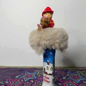 Puede incluir: Un tapón de botella decorativo con una figura de bombero, un cachorro y un acento de piel suave de color claro. El tapón de la botella tiene una escena invernal azul y blanca con el texto "Happy Holidays".