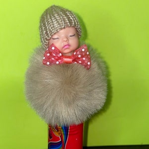 Può includere: Una piccola bambola con un cappello di lana e un fiocco rosso con pois bianchi indossa un cappotto di pelliccia marrone soffice.