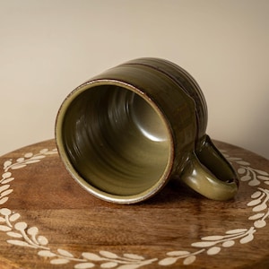 May include: An olive green ceramic mug with a handle, resting on a wooden surface. The mug has a textured exterior and a smooth interior. The wooden surface is round and decorated with a white floral pattern.