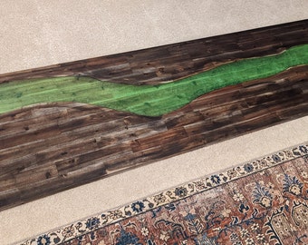 May include: A long wooden table top with a green epoxy river running through it. The wood is dark brown and has a natural grain pattern. The epoxy is smooth and shiny.