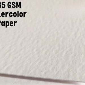 Puede incluir: Primer plano de una hoja de papel de acuarela blanco. El papel tiene una superficie texturizada y el texto "285 GSM Watercolor Paper" est&aacute; impreso en negro en la parte superior.