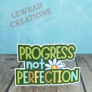 May include: A die-cut sticker featuring the text PROGRESS not PERFECTION in layered green, light blue, and tan block lettering. A white daisy with a yellow center is positioned between the words not and PERFECTION on a white border.