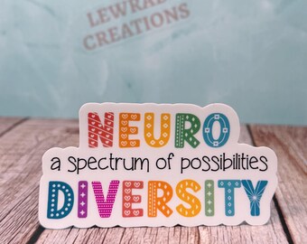 May include: A white sticker with the words "NEURO a spectrum of possibilities DIVERSITY" in colorful letters. The sticker is on a wooden surface with a light blue background. The sticker has a glossy finish.
