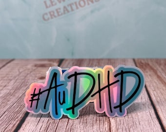 May include: A die-cut sticker featuring the text #AuDHD in bold black script lettering. The sticker has a vibrant, holographic rainbow gradient background that shifts between shades of pink, yellow, blue, and green, creating a colorful effect.