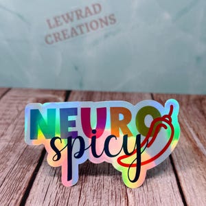 Pu&ograve; includere: Adesivo olografico con le parole "NEURO spicy" in un carattere colorato a gradiente arcobaleno, con una grafica di peperoncino rosso. L'adesivo ha una finitura lucida e si trova su una superficie di legno. Il testo "LEWRAD CREATIONS" &egrave; sullo sfondo.