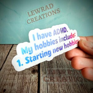 May include: A white sticker with the text "I have ADHD. My hobbies include: 1. Starting new hobbies" in blue and purple gradient lettering. The sticker is held by a hand, with the brand name "LEWRAD CREATIONS" visible in the background.