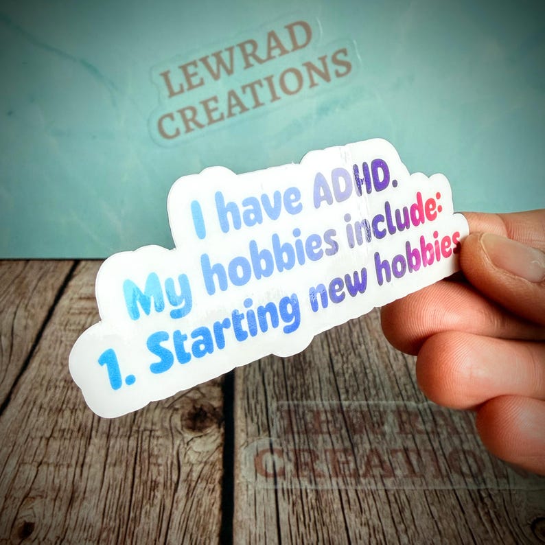 May include: A white sticker with the text "I have ADHD. My hobbies include: 1. Starting new hobbies" in blue and purple gradient lettering. The sticker is held by a hand, with the brand name "LEWRAD CREATIONS" visible in the background.