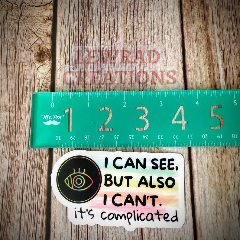 May include: A sticker with the text "I CAN SEE, BUT ALSO I CAN'T. it's complicated" and an eye graphic. A green ruler shows measurements in inches. The sticker is on a wooden surface.