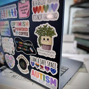May include: A laptop computer covered in colorful stickers with messages like "Be a Decent Human, It's Not That Hard", "Optimism Powered by Coffee", "See the Ability Not the Disability", "I Am a Safe Space", and "Autism Educate Accept Advocate".