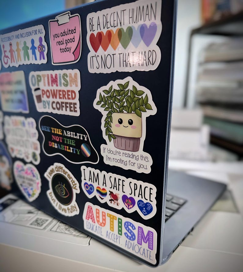 May include: A laptop computer covered in colorful stickers with messages like "Be a Decent Human, It's Not That Hard", "Optimism Powered by Coffee", "See the Ability Not the Disability", "I Am a Safe Space", and "Autism Educate Accept Advocate".