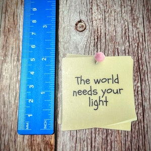 Puede incluir: Una nota adhesiva amarilla con el texto "The world needs your light" escrito en marcador negro. La nota est&aacute; fijada a una superficie de madera con un chinche rosa.