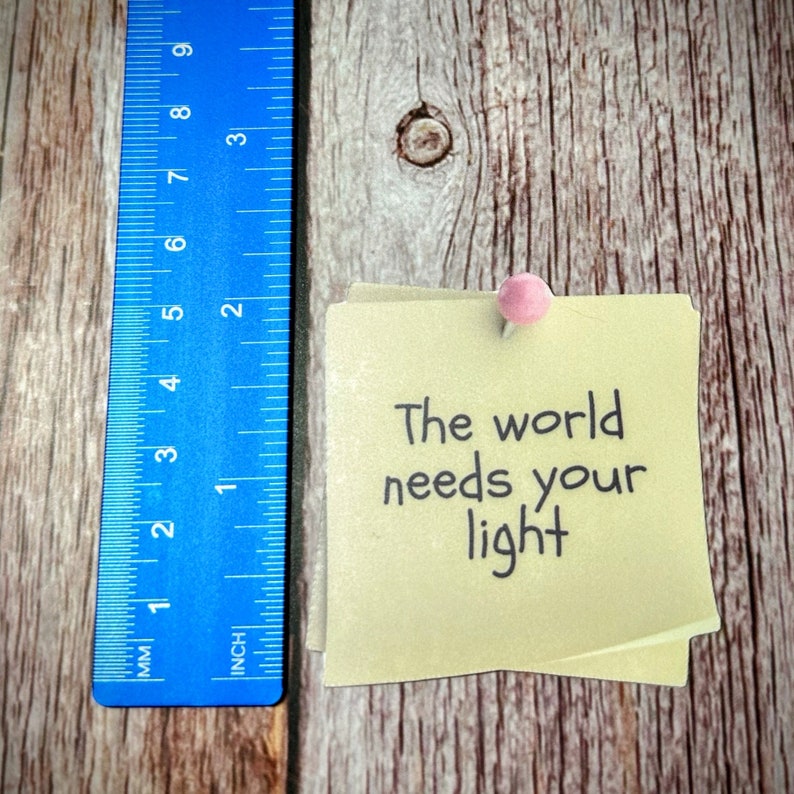 Puede incluir: Una nota adhesiva amarilla con el texto "The world needs your light" escrito en marcador negro. La nota est&aacute; fijada a una superficie de madera con un chinche rosa.