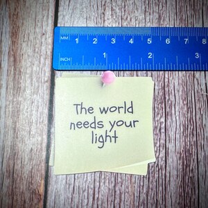Puede incluir: Una nota adhesiva amarilla con el texto "The world needs your light" escrito en marcador negro. La nota est&aacute; fijada a una superficie de madera con un chinche rosa.