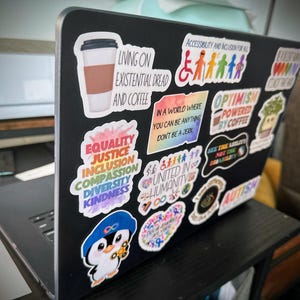 May include: A black laptop covered in colorful stickers. Stickers include phrases like "Equality, Justice, Inclusion, Compassion, Diversity, Kindness", "Living on Existential Dread and Coffee", and "Optimism Powered by Coffee". A cartoon penguin sticker is also visible.