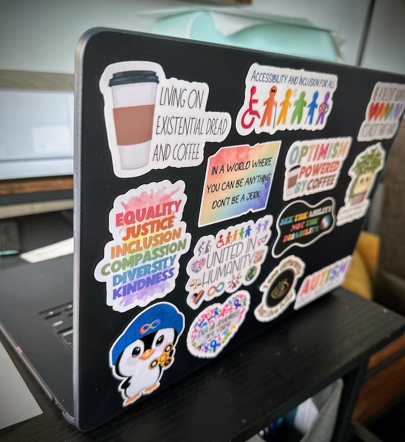 May include: A black laptop covered in colorful stickers. Stickers include phrases like "Equality, Justice, Inclusion, Compassion, Diversity, Kindness", "Living on Existential Dread and Coffee", and "Optimism Powered by Coffee". A cartoon penguin sticker is also visible.