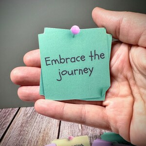 Puede incluir: Una nota adhesiva verde con el texto "Embrace the journey" escrito en marcador negro. La nota est&aacute; fijada a una mano con un chinche rosa.